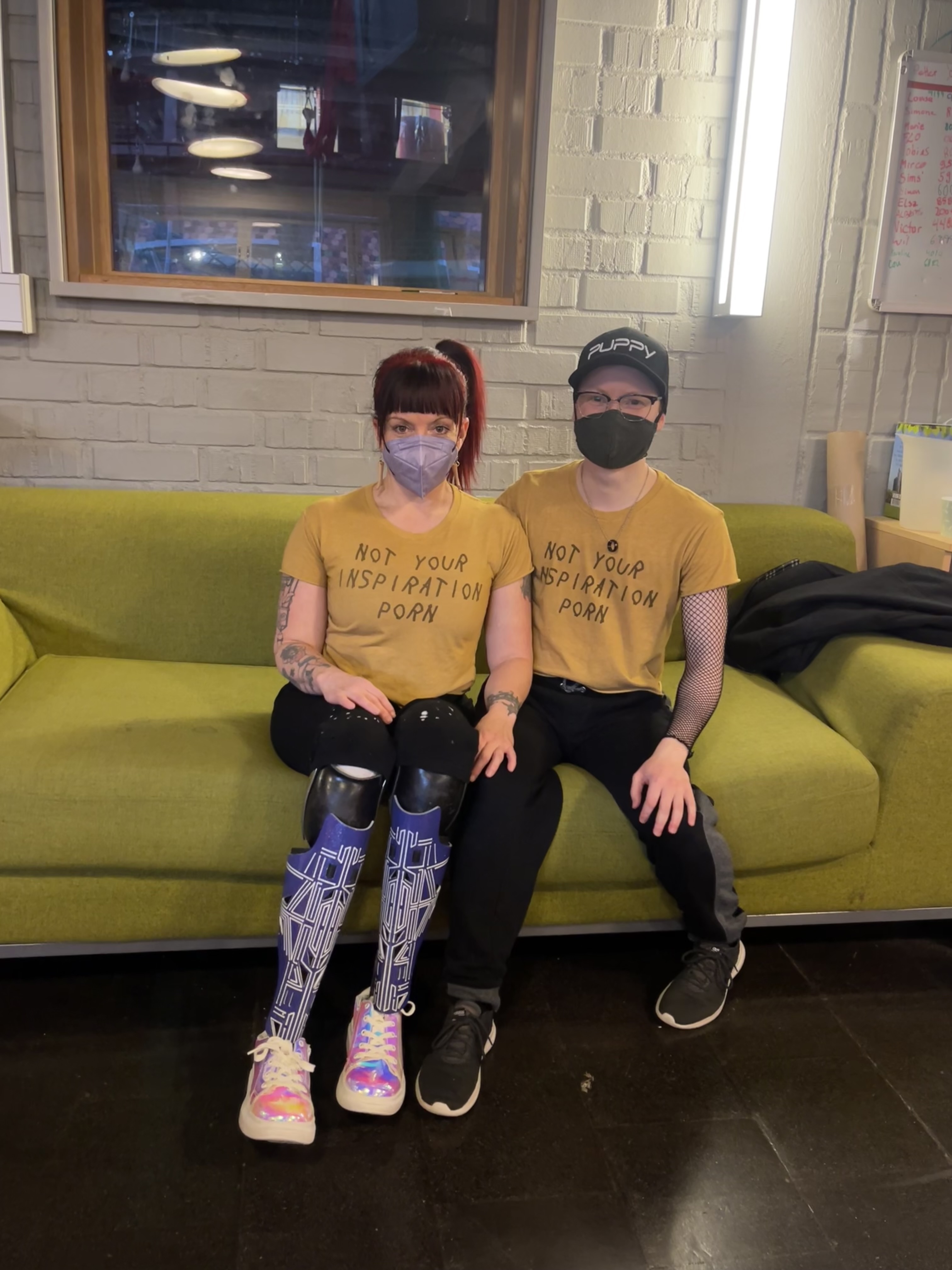 Erin and Maxime sit side by side on a couch. They wear face masks and matching shirts made by Disabled Girls Who Lift that say "Not Your Inspiration Porn".