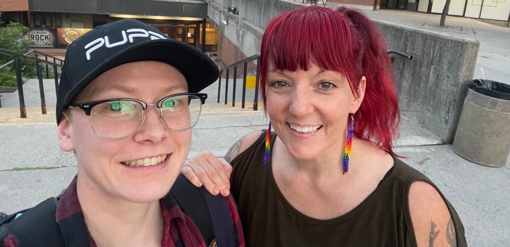 Maxime and Erin, two white Disabled humans, smile. Erin's hand rests on Maxime's shoulder. 