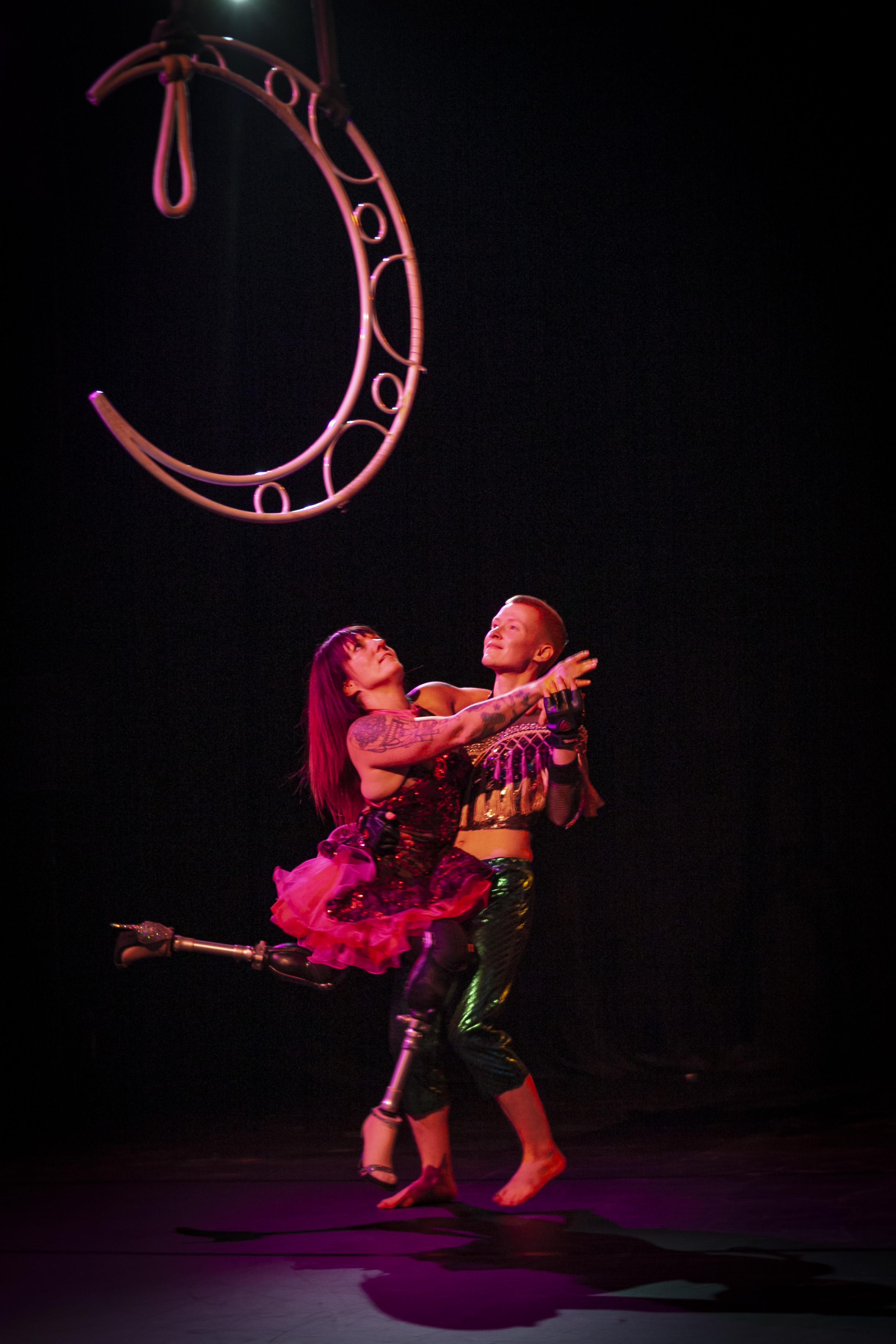 A performance photo of Erin and Maxime, dancing joyfully together and looking up at a crescent moon shaped apparatus that is suspended in the air. Erin wears high heeled prosthetic feet. 
Chris Lagesten Photography