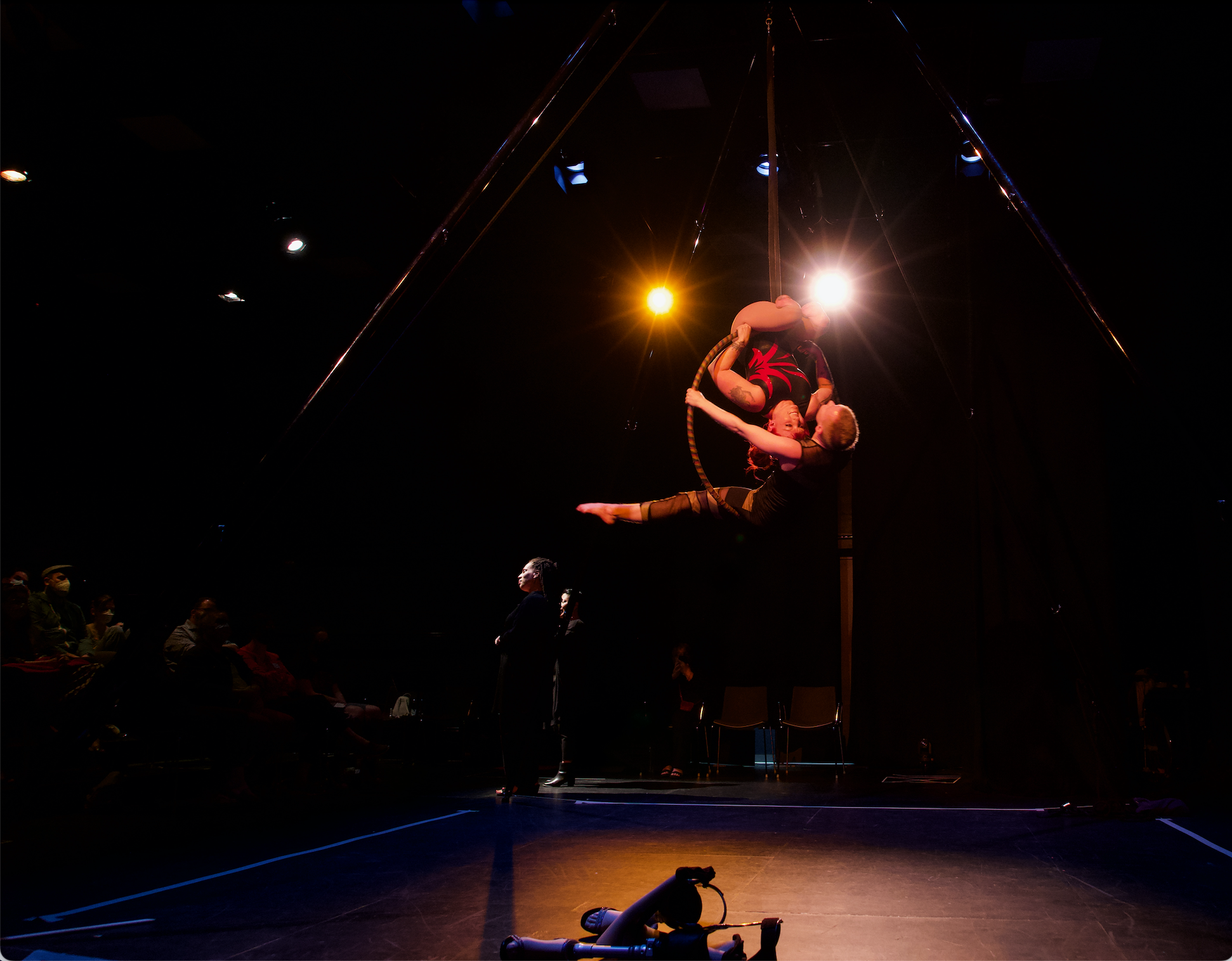 Onstage, half a story high, Erin and Maxime are on an aerial hoop: a metal hoop the size of an adult hula hoop. Erin is upside down on the top of the hoop, smiling at Maxime who sits in the hoop and leans back. On the side of the stage on the floor are Erin's prothetic legs with high heeled shoes. Photo by Jay Middaugh. FOLDA 2023.